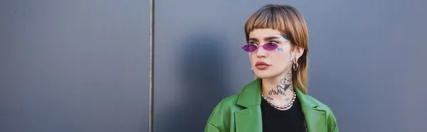 Joven mujer tatuada en gafas y chaqueta verde mirando al aire libre, pancarta - foto de stock