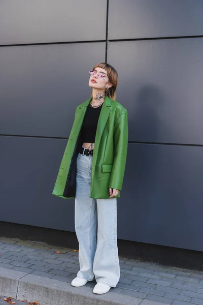 Visão comprimento total da mulher tatuada em jaqueta de couro verde e jeans de pé perto da parede na rua da cidade — Fotografia de Stock