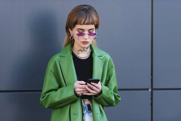 Mensajería mujer tatuada de moda en el teléfono inteligente cerca de la pared gris al aire libre - foto de stock