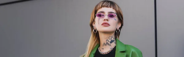 Stylish tattooed woman wearing eyeglasses and looking at camera near grey wall, banner — Stock Photo