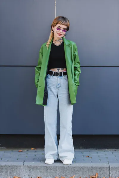 Donna tatuata in jeans in piedi con le mani in tasche di giacca verde vicino alla parete grigia — Foto stock