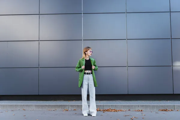 Full length view of tattooed woman in stylish autumn clothes looking away near wall on city street — Stock Photo