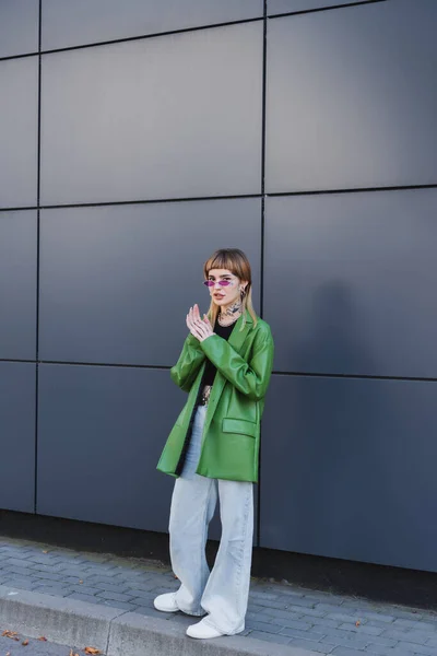 Femme tatouée dans des vêtements d'automne élégants et des lunettes debout près du mur gris — Photo de stock