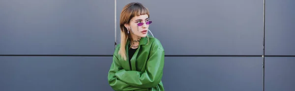 Mujer tatuada en gafas con estilo y chaqueta de cuero verde mirando hacia otro lado cerca de la pared gris, pancarta - foto de stock