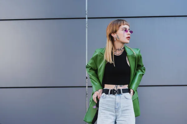 Mujer tatuada en chaqueta verde y jeans de pie con las manos en las caderas cerca de la pared gris - foto de stock