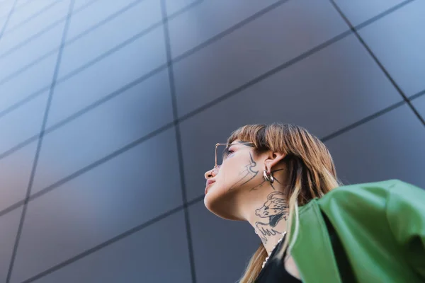 Low angle view of stylish tattooed woman near high grey wall on urban street — Stock Photo