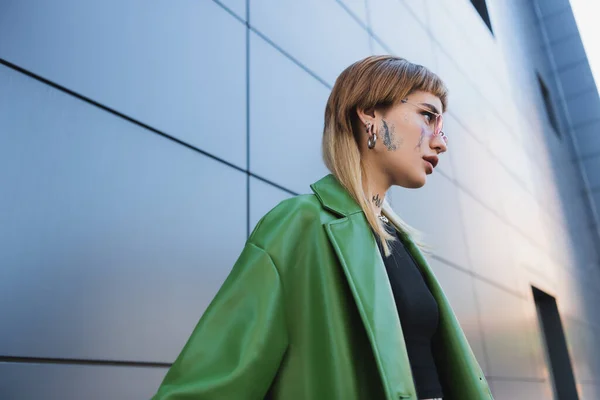 Tattooed woman with piercing wearing green leather jacket near grey wall outdoors — Stock Photo