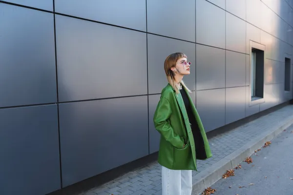 Mujer joven y elegante en chaqueta de cuero verde de pie con la mano en el bolsillo cerca de la pared gris - foto de stock