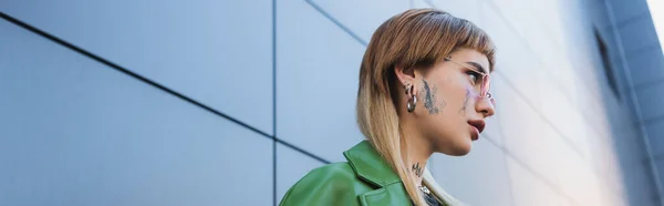 Vista de ángulo bajo de la mujer joven con tatuaje y piercing cerca de la pared gris al aire libre, pancarta - foto de stock