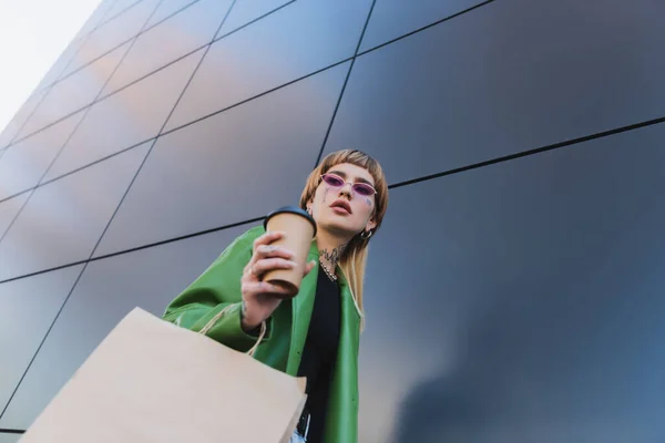 Vista de ángulo bajo de la mujer tatuada con estilo con café para ir y bolsa de compras cerca de la pared gris - foto de stock