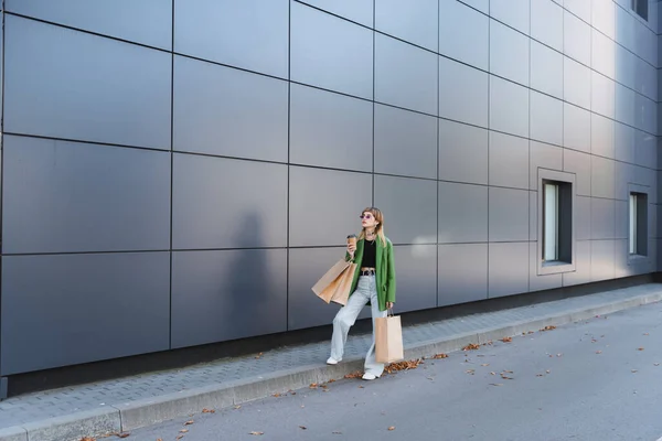 Mujer joven y elegante con bolsas de compras y café para ir de pie cerca de la pared gris alto - foto de stock
