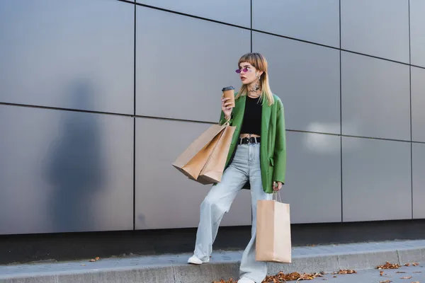 Elegante donna tatuata in giacca verde e jeans in piedi con borse della spesa e tazza di carta all'aperto — Foto stock