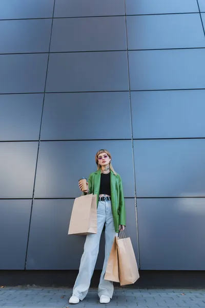 Модная татуированная женщина в джинсах, стоящая рядом с серой стеной с бумажной чашкой и пакетами для покупок — стоковое фото