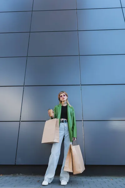 Trendy tattooed woman in jeans standing near grey wall with paper cup and shopping bags — Stock Photo