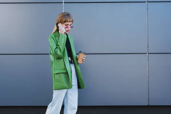 Mujer de moda en chaqueta de cuero verde sosteniendo taza de papel mientras habla en el teléfono móvil al aire libre - foto de stock