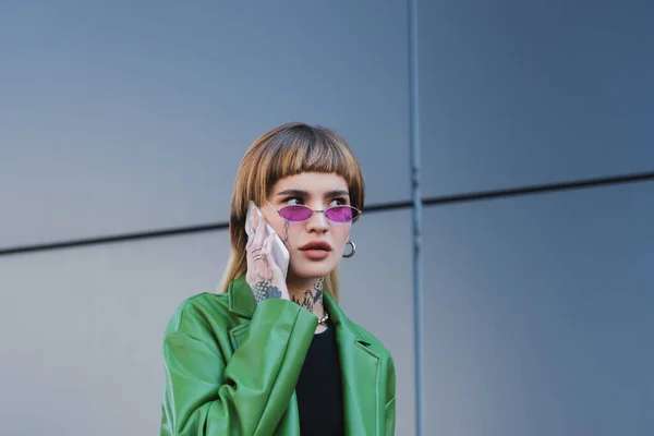 Joven mujer tatuada en chaqueta verde llamando al celular en la calle de la ciudad - foto de stock