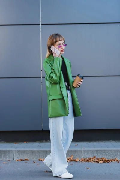 Ganzkörperansicht einer stilvollen Frau mit Imbissgetränk, die im Freien auf dem Smartphone spricht — Stockfoto