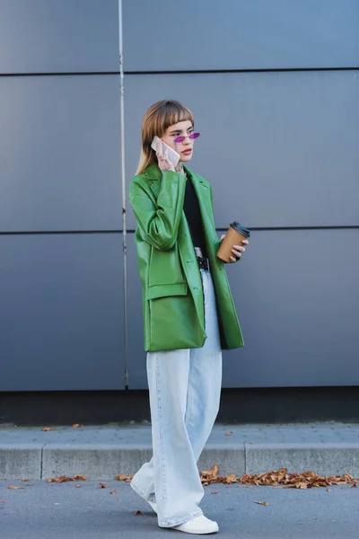 Full length view of stylish woman with takeaway drink talking on smartphone outdoors — Stock Photo