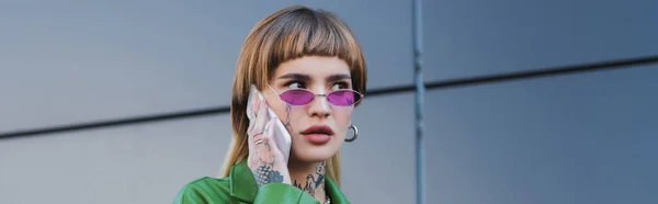 Молода татуйована жінка в окулярах говорить на мобільному телефоні на вулиці міста, банер — стокове фото