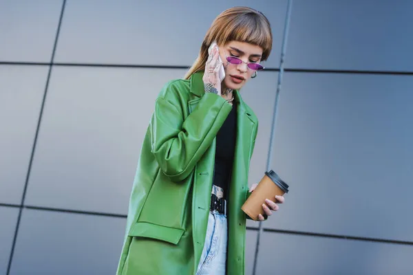 Donna tatuata in giacca di pelle verde che tiene il caffè per andare a chiamare il cellulare all'aperto — Foto stock