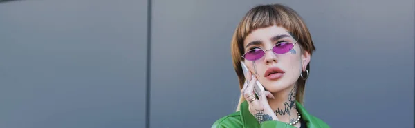 Tätowierte Frau mit stylischer Brille, die in der Nähe der grauen Wand mit dem Handy spricht, Banner — Stockfoto