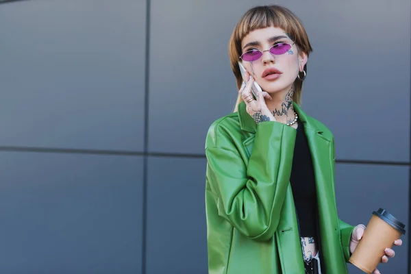 Mujer elegante y tatuada con bebida para llevar hablando en un teléfono inteligente cerca de la pared gris - foto de stock