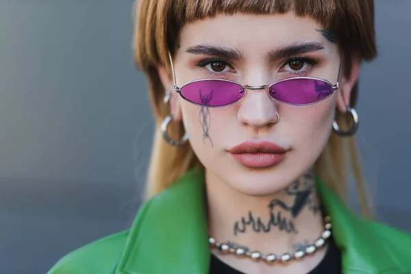 Portrait de femme tatouée dans des lunettes à la mode regardant la caméra à l'extérieur — Photo de stock