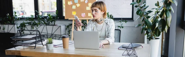 Молода і стильна бізнес-леді, використовуючи мобільний телефон під час роботи біля ноутбука в офісі, банер — стокове фото