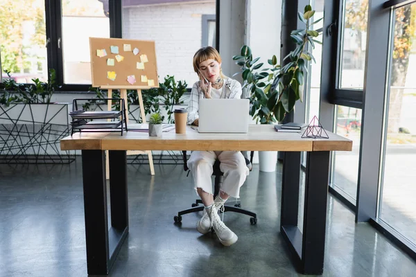 Young tattooed businesswoman talking on mobile phone while working at laptop in office — Stock Photo