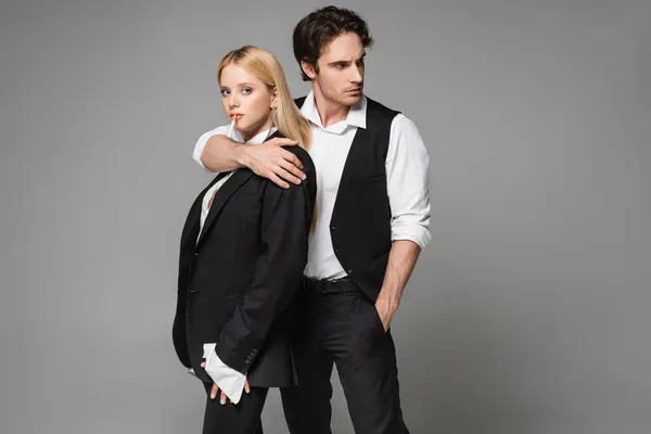 Blonde woman with cigarette looking at camera while stylish man embracing her isolated on grey — Stock Photo