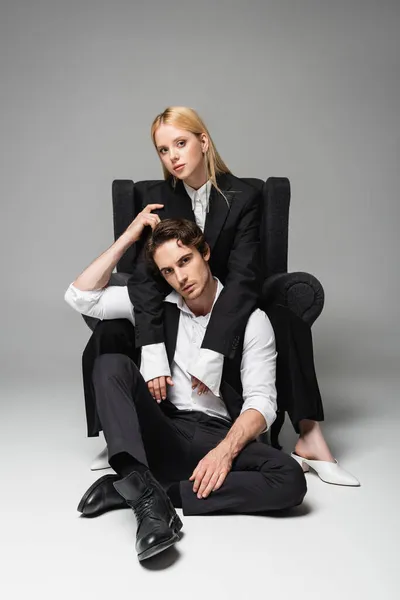 Blonde Frau im Sessel umarmt eleganten Mann, der auf dem Boden auf grau sitzt — Stockfoto