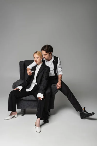 Stylish man lightening cigarette of blonde woman while sitting in armchair on grey — Stock Photo