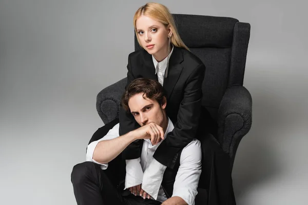 Blonde woman in armchair looking at camera while embracing stylish man sitting on grey — Stock Photo