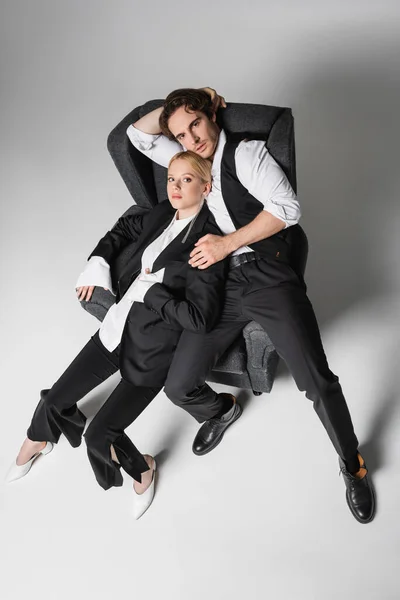 Vista de ángulo alto de elegante pareja en ropa blanca y negra posando en sillón sobre gris - foto de stock