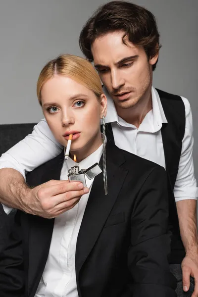 Young blonde woman looking at camera while stylish man lightening her cigarette isolated on grey — Stock Photo