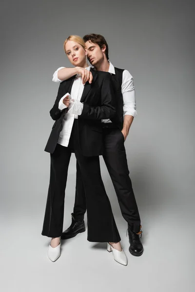 Visão de comprimento total de homem elegante abraçando mulher loira enquanto está de pé com a mão no bolso em cinza — Fotografia de Stock