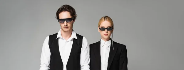 Front view of young couple in elegant clothes and dark sunglasses standing isolated on grey, banner — Stock Photo