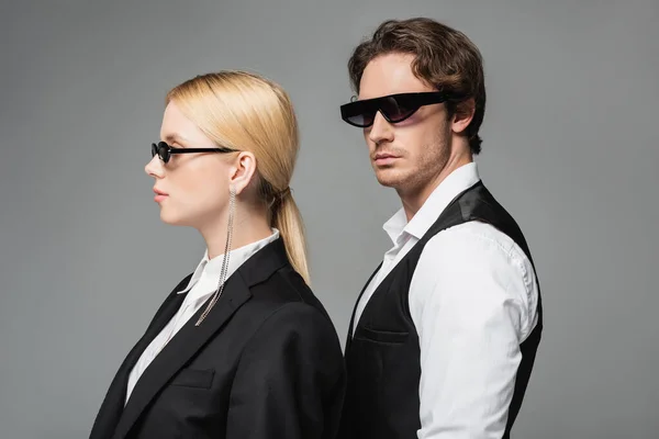 Femme blonde et brune homme aux lunettes de soleil sombres et aux vêtements élégants posant isolé sur gris — Photo de stock