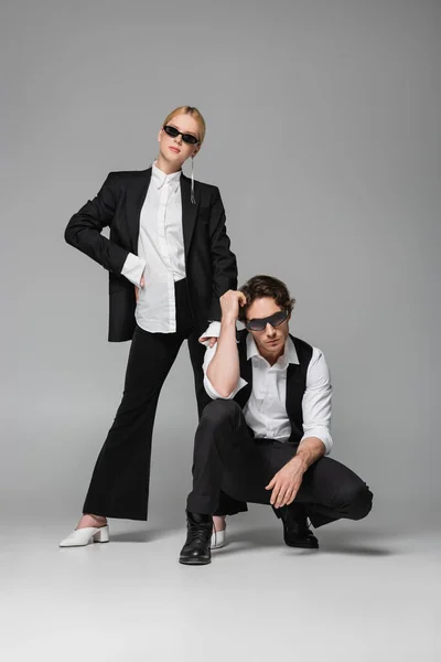 Blonde woman in black suit standing with hand on hip near elegant man posing on grey — Stock Photo