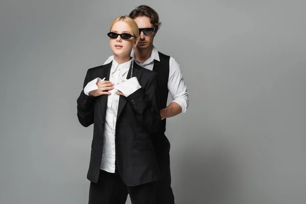 Pareja con estilo en gafas de sol oscuras y ropa en blanco y negro posando aislada en gris - foto de stock