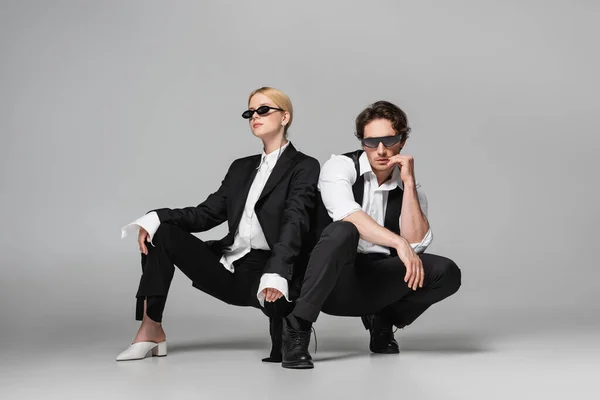 Elegant couple in black suits and dark sunglasses posing on grey — Stock Photo