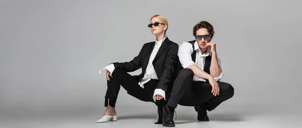 Pareja joven en trajes elegantes y gafas de sol oscuras posando en gris, pancarta - foto de stock