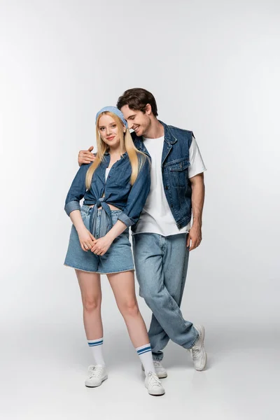 Homem feliz em roupas jeans abraçando namorada bonita e elegante olhando para a câmera no branco — Fotografia de Stock