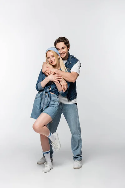 Feliz joven pareja en ropa de mezclilla mirando a la cámara mientras se abraza en blanco - foto de stock