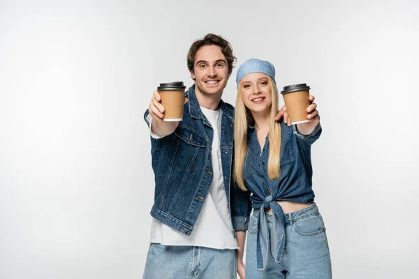 Cheerful couple in stylish denim clothing holding disposable cups in outstretched hands isolated on white — Stock Photo