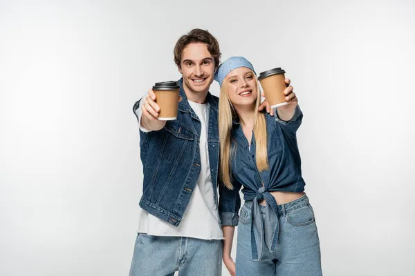 Couple souriant dans des vêtements en denim à la mode montrant des tasses en papier isolé sur blanc — Photo de stock