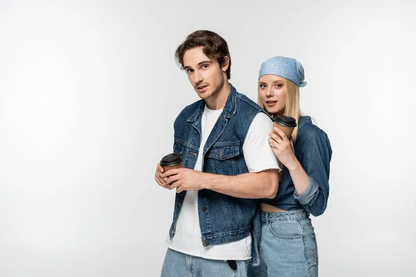 Feliz jovem casal em roupas jeans segurando copos de papel enquanto olha para a câmera isolada no branco — Fotografia de Stock