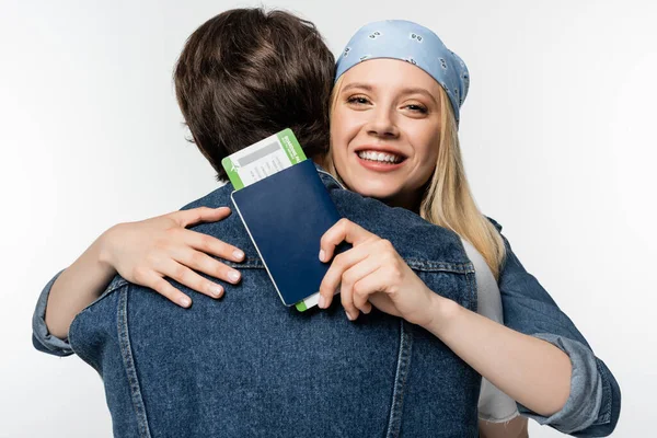 Mujer alegre en diadema sosteniendo documentos de viaje mientras abraza novio aislado en blanco - foto de stock