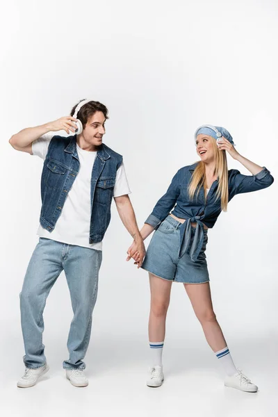 Couple joyeux en denim vêtements et écouteurs tenant la main tout en écoutant de la musique sur blanc — Photo de stock