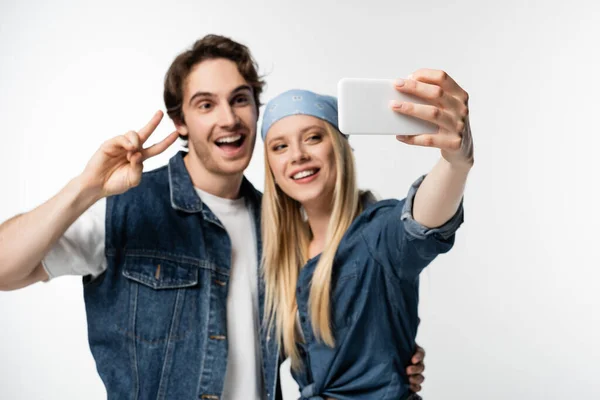 Mujer feliz tomando selfie con novio mostrando signo de paz en fondo borroso aislado en blanco - foto de stock